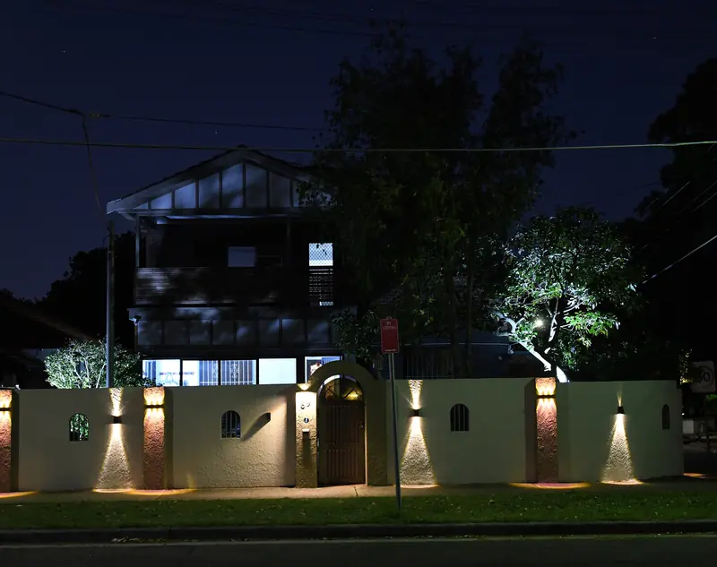 A garden wall and trees lit by uplights in front of a house at night