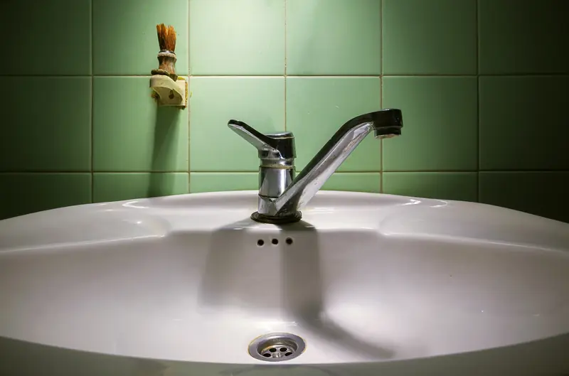 A chrome faucet over a white ceramic sink in front of green tiles